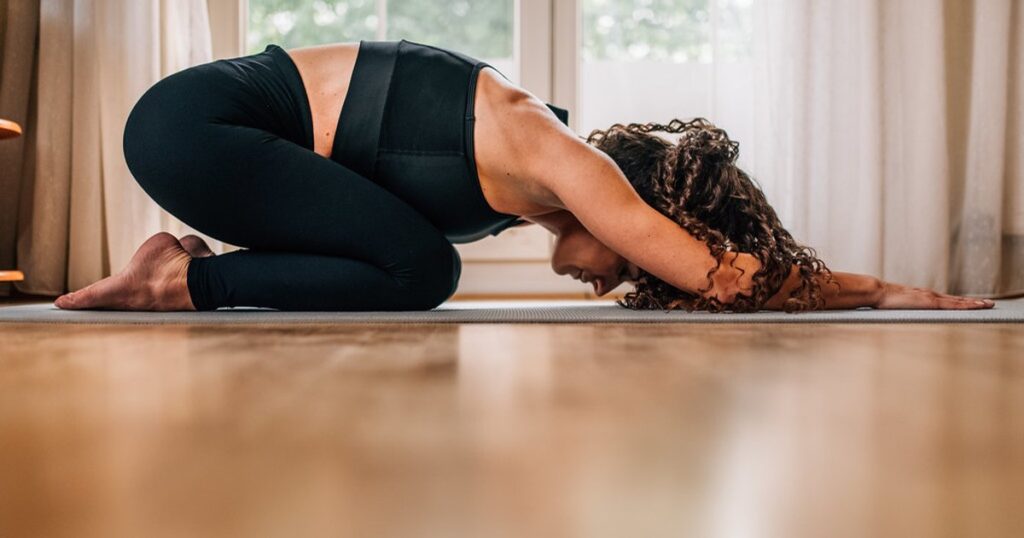 A woman in extended puppy pose stretches forward with her arms and chest lowered toward a yoga mat on the floor.